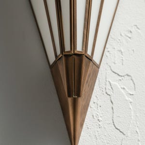 May include: A wall sconce featuring a triangular, pointed base made of aged bronze-toned metal. The upper section consists of white, opaque glass panels framed by thin, vertical bronze-toned metal strips, creating a geometric, fan-like structure.