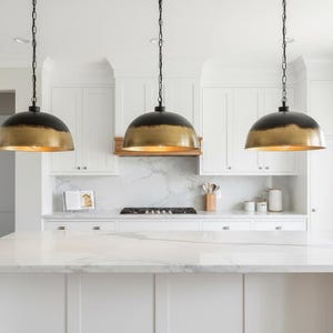 Peut inclure: Trois suspensions en forme de dôme avec un extérieur noir mat et un intérieur doré métallique. Chaque luminaire est suspendu par une chaîne noire au-dessus d'un îlot de cuisine blanc avec un comptoir en marbre dans une cuisine lumineuse.