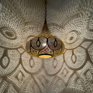 May include: A gold Moroccan pendant light with intricate cut-out designs casts a beautiful pattern of light on the ceiling. The light is hanging from a chain.