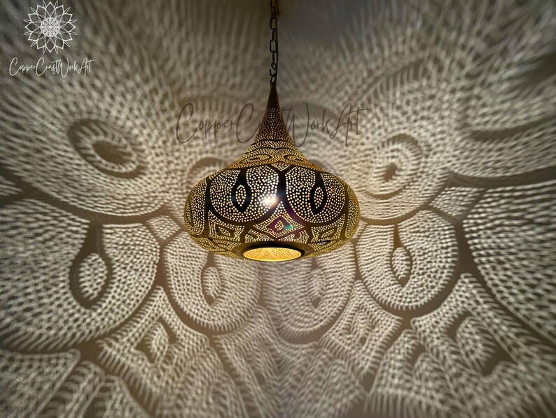 May include: A gold Moroccan pendant light with intricate cut-out designs casts a beautiful pattern of light on the ceiling. The light is hanging from a chain.
