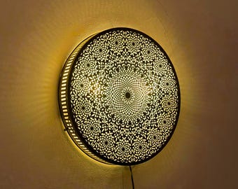 May include: Round, decorative wall sconce with a detailed, openwork design. The metal fixture features a circular pattern of floral and geometric shapes, emitting a warm, inviting glow. The sconce is mounted on a neutral-colored wall.