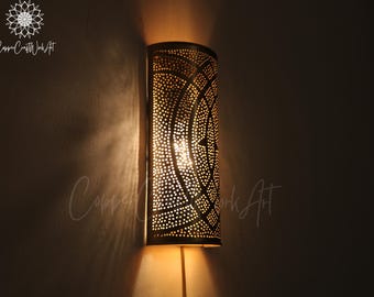 May include: A gold metal wall sconce with a geometric pattern. The sconce is lit and casting a warm glow on the wall.