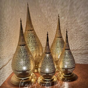 Op de afbeelding: Vijf goudkleurige, sierlijke metalen lantaarns met ingewikkelde uitgesneden patronen. Elke lantaarn heeft een druppelvorm, een ronde basis en een puntige top. De lantaarns zijn van binnenuit verlicht en werpen patronen op de achtergrond.