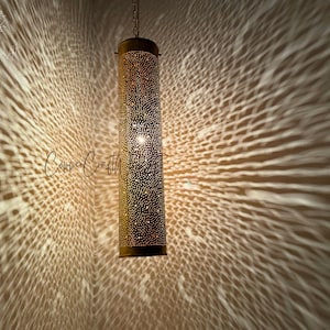 May include: A gold metal pendant light fixture with a cylindrical shade. The shade is covered in small holes, creating a pattern of light and shadow when illuminated.