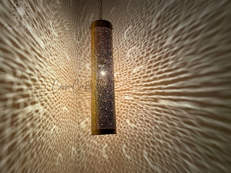 May include: A gold metal pendant light fixture with a cylindrical shade. The shade is covered in small holes, creating a pattern of light and shadow when illuminated.