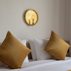 May include: A gold circular wall sconce with a lit candle mounted above a bed featuring two white pillows and two mustard yellow square throw pillows. The wall sconce has a reflective metallic finish and holds a single pillar candle.