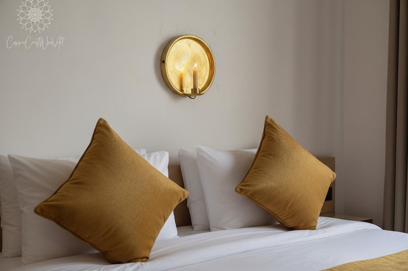 May include: A gold circular wall sconce with a lit candle mounted above a bed featuring two white pillows and two mustard yellow square throw pillows. The wall sconce has a reflective metallic finish and holds a single pillar candle.
