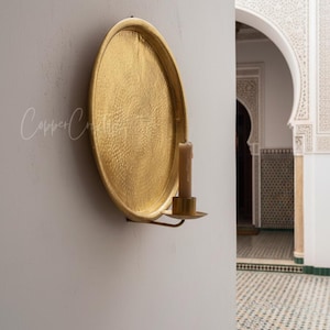 May include: A circular gold-colored metal wall sconce featuring a large, hammered-texture round backplate and a small cylindrical candle holder attached to the bottom. A single beige pillar candle sits inside the holder on the metal arm.