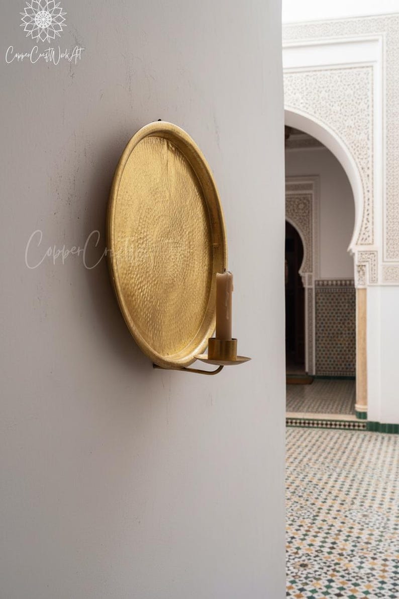 May include: A circular gold-colored metal wall sconce featuring a large, hammered-texture round backplate and a small cylindrical candle holder attached to the bottom. A single beige pillar candle sits inside the holder on the metal arm.