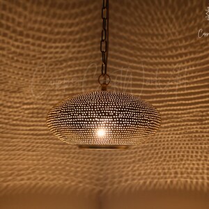 May include: A gold-toned metal pendant light with a rounded, flattened shape featuring a dense pattern of small, circular perforations. The fixture is suspended by a dark metal chain and emits a warm light that casts a textured shadow pattern.