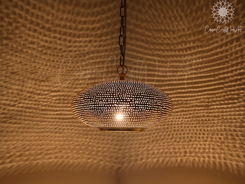 May include: A gold-toned metal pendant light with a rounded, flattened shape featuring a dense pattern of small, circular perforations. The fixture is suspended by a dark metal chain and emits a warm light that casts a textured shadow pattern.