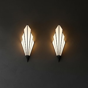 May include: Two wall sconces featuring a fan-shaped design with black metal frames and white translucent panels. Each light fixture emits a warm glow from within, showcasing a pointed base and a symmetrical, multi-tiered geometric structure.