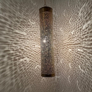 May include: A gold metal pendant light fixture with a cylindrical shade. The shade is covered in small holes, creating a pattern of light and shadow when illuminated.