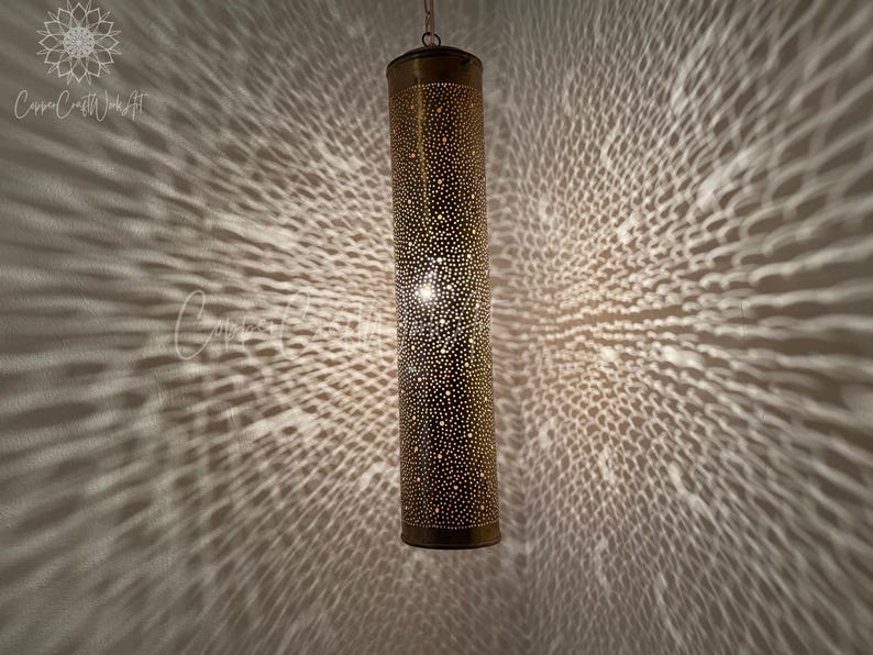 May include: A gold metal pendant light fixture with a cylindrical shade. The shade is covered in small holes, creating a pattern of light and shadow when illuminated.