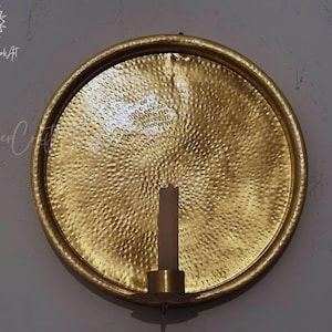 May include: A circular gold-colored metal wall sconce featuring a hammered texture across its surface. The piece includes a central cylindrical candle holder at the bottom, designed to hold a single pillar candle. The text CopperCraftWorkArt is visible.
