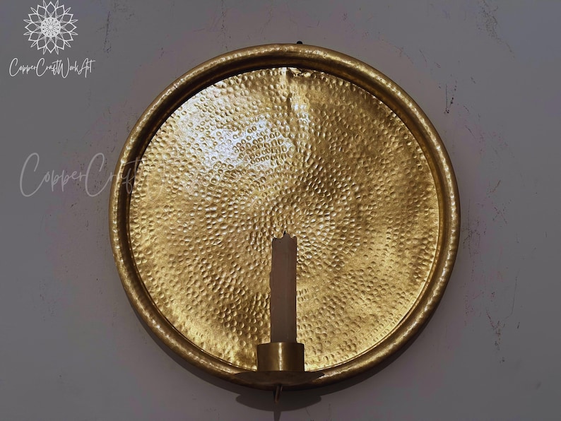 May include: A circular gold-colored metal wall sconce featuring a hammered texture across its surface. The piece includes a central cylindrical candle holder at the bottom, designed to hold a single pillar candle. The text CopperCraftWorkArt is visible.