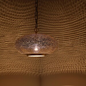 May include: A gold-toned metal pendant light with a rounded, flattened shape featuring a dense pattern of small, circular perforations. The fixture is suspended by a dark metal chain and emits a warm glow, casting intricate light patterns.