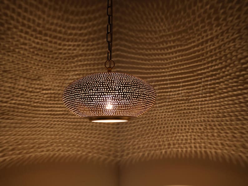 May include: A gold-toned metal pendant light with a rounded, flattened shape featuring a dense pattern of small, circular perforations. The fixture is suspended by a dark metal chain and emits a warm glow, casting intricate light patterns.