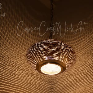 May include: A round, metallic pendant light fixture with a dark, perforated surface that casts a complex, web-like shadow pattern. The lamp hangs from a chain and features a central circular opening emitting warm light. Text reads CopperCraftWorkArt.