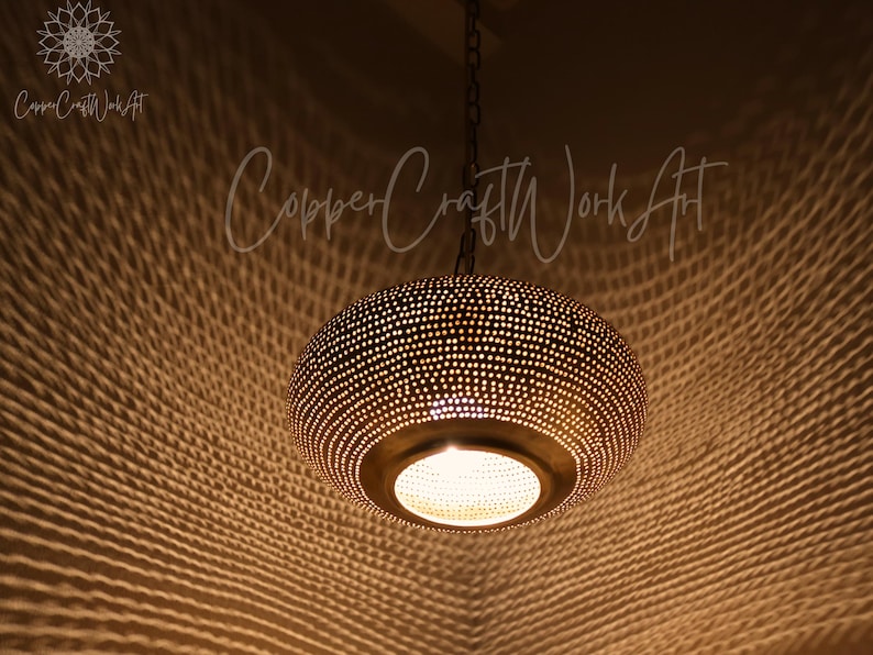 May include: A round, metallic pendant light fixture with a dark, perforated surface that casts a complex, web-like shadow pattern. The lamp hangs from a chain and features a central circular opening emitting warm light. Text reads CopperCraftWorkArt.