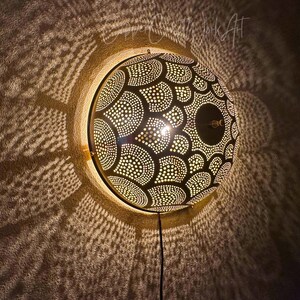 May include: A round, black wall lamp with a patterned design of white dots and semi-circles. The lamp casts intricate light patterns on the wall. A black cord extends from the bottom.