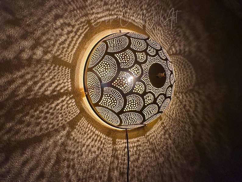 May include: A round, black wall lamp with a patterned design of white dots and semi-circles. The lamp casts intricate light patterns on the wall. A black cord extends from the bottom.