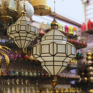Peut inclure: Suspensions lumineuses géométriques avec des panneaux de verre blanc et des cadres en laiton. L'arrière-plan présente des étagères remplies d'objets décoratifs variés, créant une ambiance de magasin.