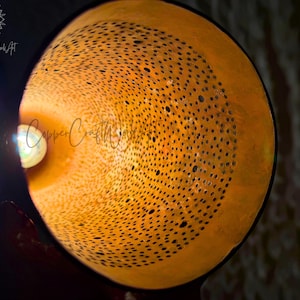 May include: A close-up of a copper lampshade with a circular pattern of holes. The lampshade is lit from the inside, casting a warm glow.