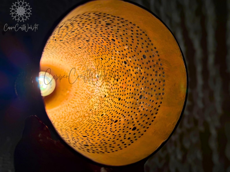 May include: A close-up of a copper lampshade with a circular pattern of holes. The lampshade is lit from the inside, casting a warm glow.