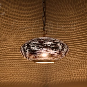 May include: A metallic pendant light fixture with a rounded, flattened shape and a perforated surface pattern. The fixture is suspended by a dark chain and emits a warm, glowing light through the small holes, casting a textured shadow pattern.