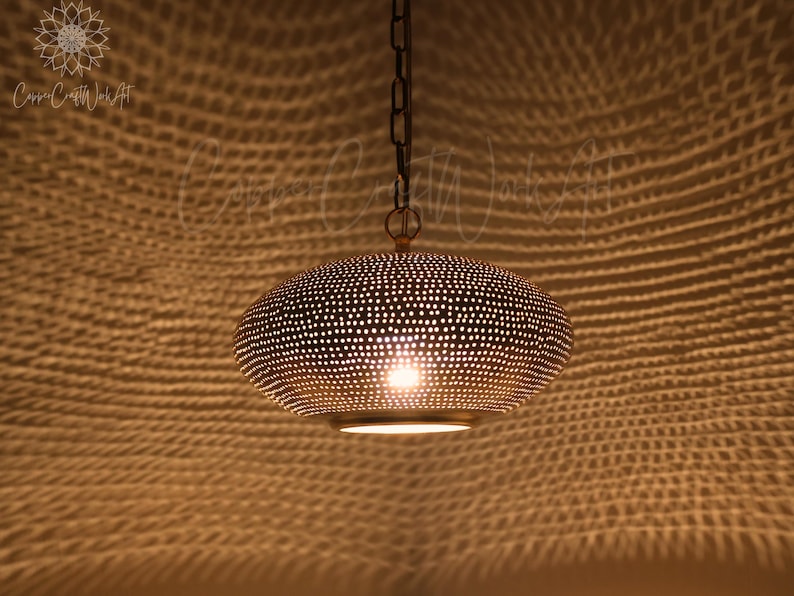 May include: A metallic pendant light fixture with a rounded, flattened shape and a perforated surface pattern. The fixture is suspended by a dark chain and emits a warm, glowing light through the small holes, casting a textured shadow pattern.