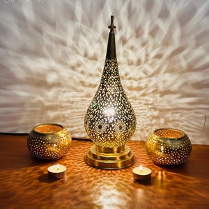 May include: A gold metal table lamp with a decorative, intricate, cut-out design. The lamp is on a wooden table with two smaller gold metal candle holders and two white tea lights.