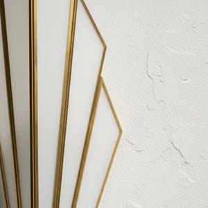 May include: A wall sconce featuring a series of white glass panels framed in thin gold metal strips. The design consists of multiple vertical and angled segments arranged in a fan-like pattern, creating a geometric structure with clean lines.