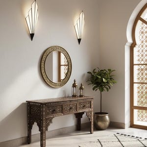 May include: A dark wood console table with intricate white inlay patterns and three drawers, positioned below a round gold-framed mirror. Two geometric wall sconces with white panels are mounted above, and two metal lanterns sit on the table surface.