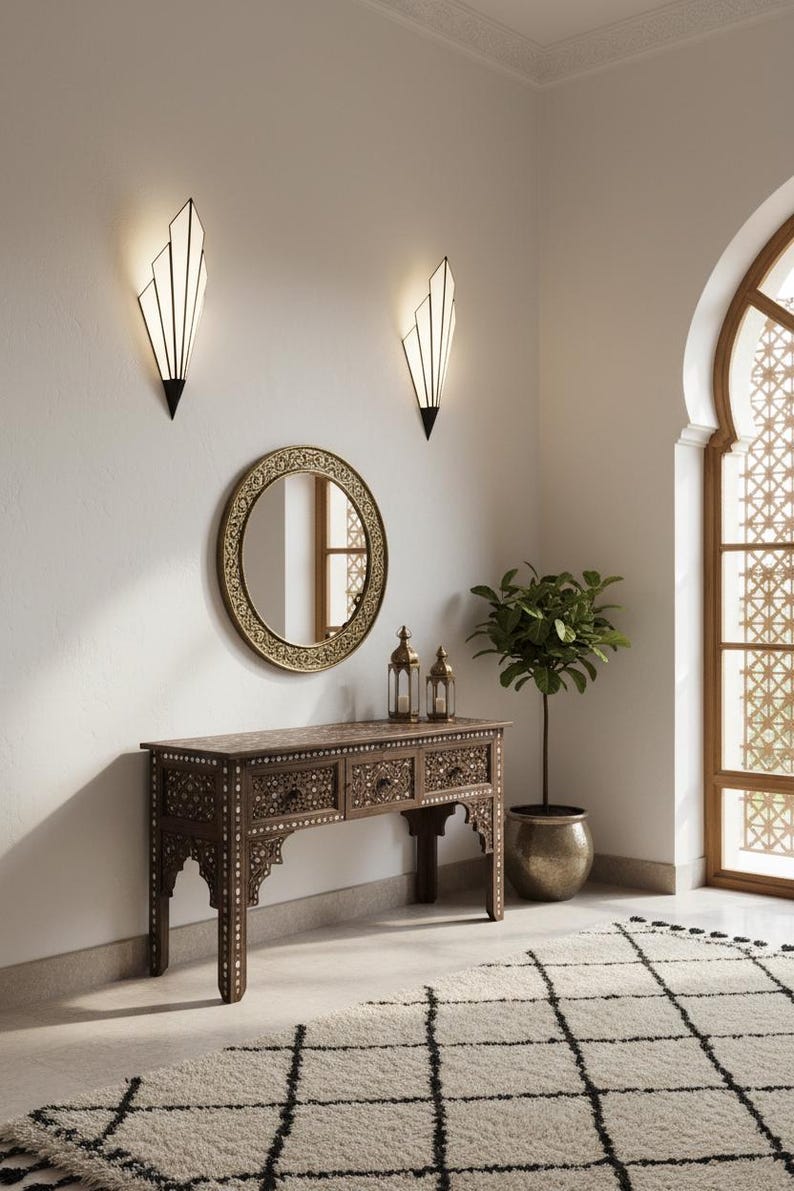 May include: A dark wood console table with intricate white inlay patterns and three drawers, positioned below a round gold-framed mirror. Two geometric wall sconces with white panels are mounted above, and two metal lanterns sit on the table surface.