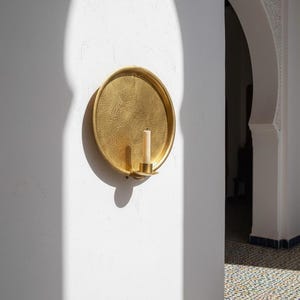 May include: A circular gold-toned metal wall sconce featuring a hammered texture on the surface. The sconce includes a small cylindrical holder at the bottom center, which contains a single off-white pillar candle. The metal has a reflective finish.