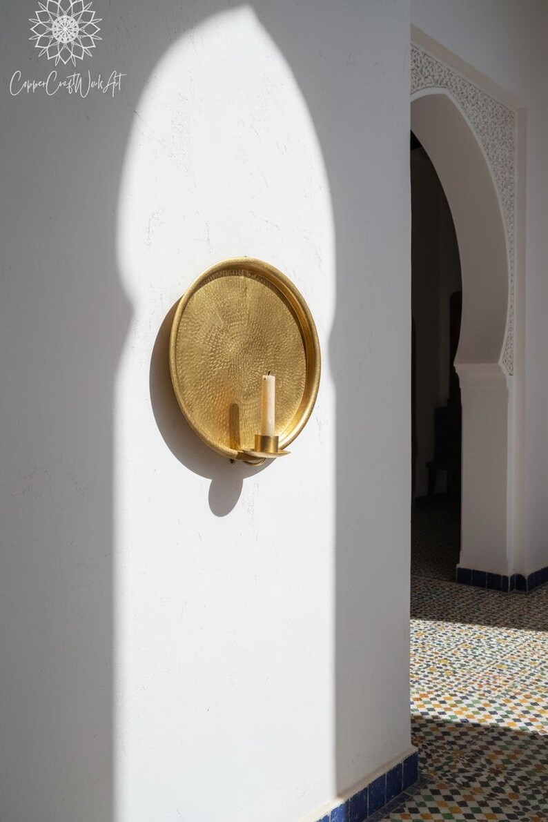 May include: A circular gold-toned metal wall sconce featuring a hammered texture on the surface. The sconce includes a small cylindrical holder at the bottom center, which contains a single off-white pillar candle. The metal has a reflective finish.