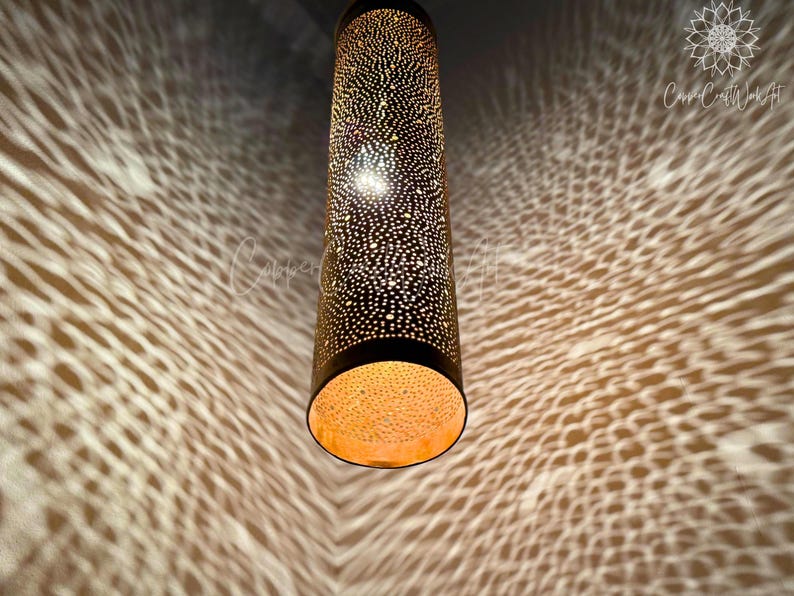 May include: A gold metal lampshade with a perforated pattern casts a decorative shadow on the ceiling. The lampshade is cylindrical and has a warm, inviting glow.
