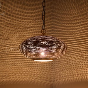 May include: A metallic pendant light fixture with a rounded, perforated dome shape that casts intricate, web-like shadows on surrounding surfaces. The lamp is suspended by a dark metal chain and emits a warm, focused glow from its center.