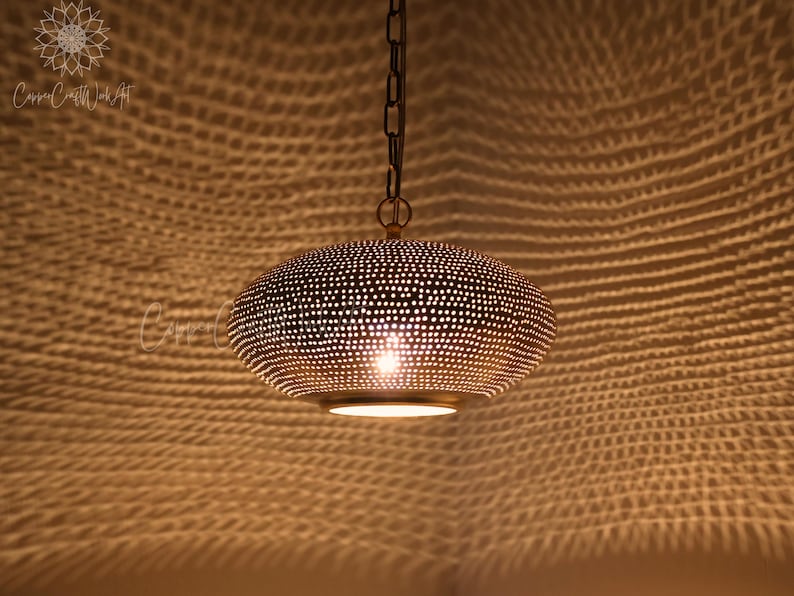 May include: A metallic pendant light fixture with a rounded, perforated dome shape that casts intricate, web-like shadows on surrounding surfaces. The lamp is suspended by a dark metal chain and emits a warm, focused glow from its center.