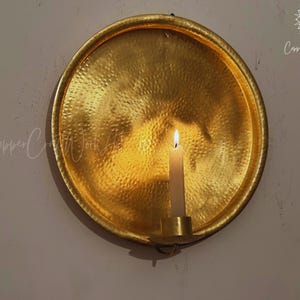 May include: A circular, gold-toned metal wall sconce featuring a hammered texture. The piece includes a central candle holder with a lit white taper candle. The text CopperCraftWorkArt is visible in the top right corner and center left.