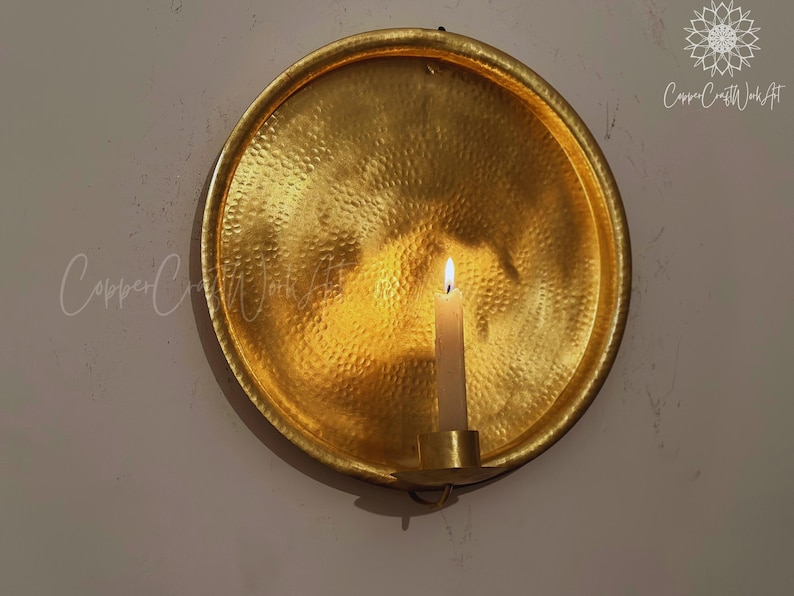 May include: A circular, gold-toned metal wall sconce featuring a hammered texture. The piece includes a central candle holder with a lit white taper candle. The text CopperCraftWorkArt is visible in the top right corner and center left.