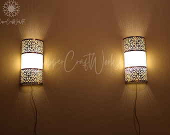 May include: Two gold metal wall sconces with white frosted glass shades and intricate cut-out designs. The sconces are mounted on a beige wall and are illuminated.