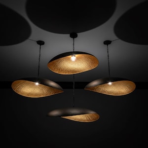 Op de afbeelding: Vier hanglampen met zwarte, getextureerde buitenschalen en reflecterende, gehamerde goudkleurige interieurs. Elke lamp hangt aan een dunne zwarte ketting en heeft een centrale gloeilamp, wat zorgt voor een warme gloed tegen de donkere achtergrond.
