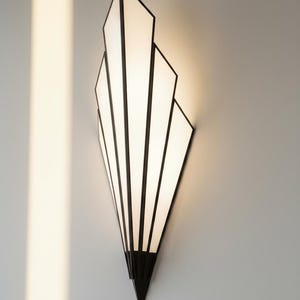 May include: A wall sconce featuring a fan-shaped design with a black metal frame and white translucent panels. The fixture emits a soft, warm glow from behind the panels, creating a pointed, geometric silhouette that tapers to a sharp bottom point.