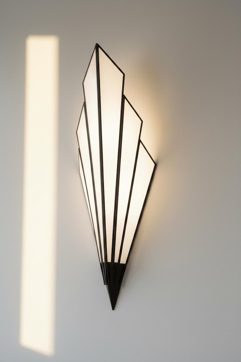 May include: A wall sconce featuring a fan-shaped design with a black metal frame and white translucent panels. The fixture emits a soft, warm glow from behind the panels, creating a pointed, geometric silhouette that tapers to a sharp bottom point.