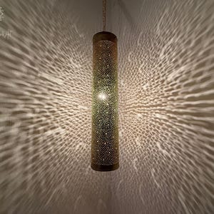 May include: A gold metal hanging lamp with a cylindrical shape and a patterned design. The lamp casts a geometric shadow on the wall.