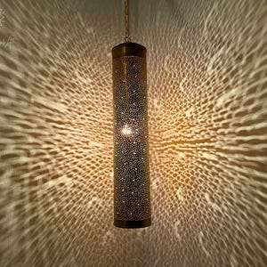May include: A gold metal pendant light fixture with a cylindrical shade. The shade is covered in small holes, creating a pattern of light and shadow when lit.