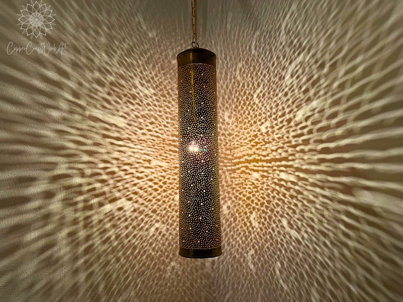 May include: A gold metal pendant light fixture with a cylindrical shade. The shade is covered in small holes, creating a pattern of light and shadow when lit.