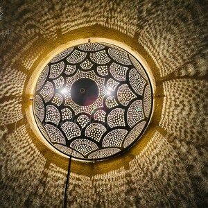 May include: A round, black and gold metal wall lamp with a detailed, patterned design. The lamp casts intricate light patterns on the surrounding wall. The lamp is mounted on a wall and has a power cord.